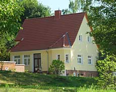 Haus 13 - ein kleines, gelbes Haus mit rotem Satteldach. Ringsherum Bäume.