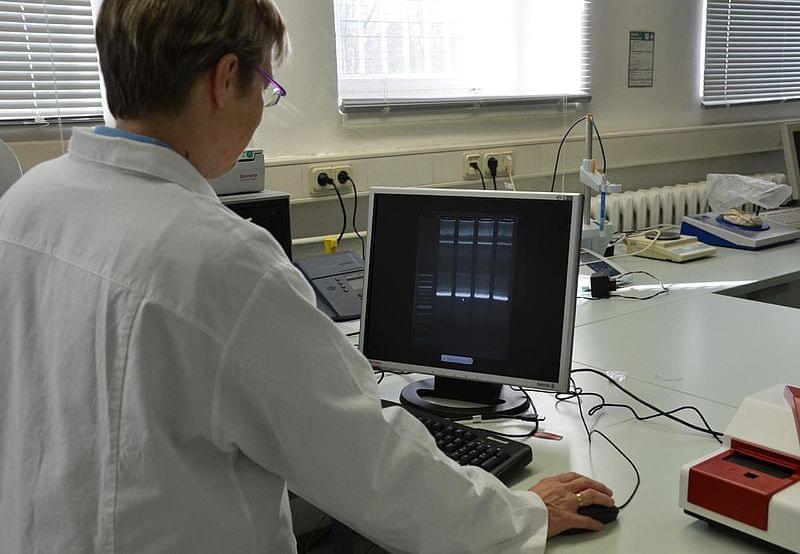 Eine Person in einem weißen Laborkittel sitzt an einem Computer in einem Labor und beobachtet den Bildschirm, auf dem wissenschaftliche Daten oder Diagramme angezeigt werden. Elektronische Geräte und Laborzubehör sind im Hintergrund sichtbar.