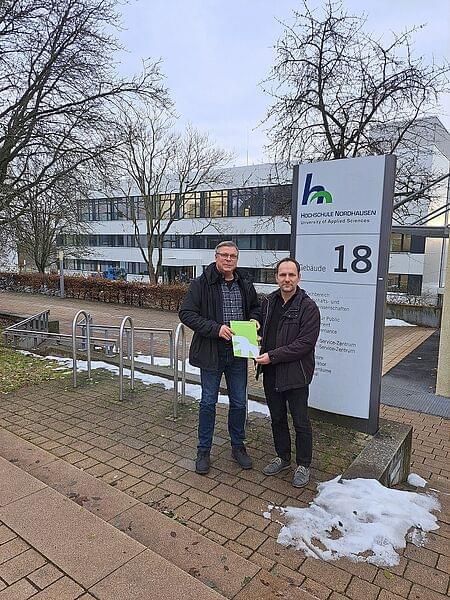 Heiko Lange and Prof Dr Mario Schölzel stand next to each other in front of the information board for House 18, holding a green HSN document folder. In the background is House 18, with remnants of snow on the ground and bare trees.