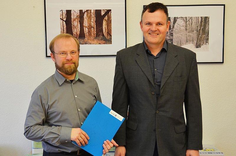 Prof Dr Hinz and Prof Dr Wagner are standing next to each other. Prof Hinz is holding a blue folder. Framed photographs hang on the wall in the background.