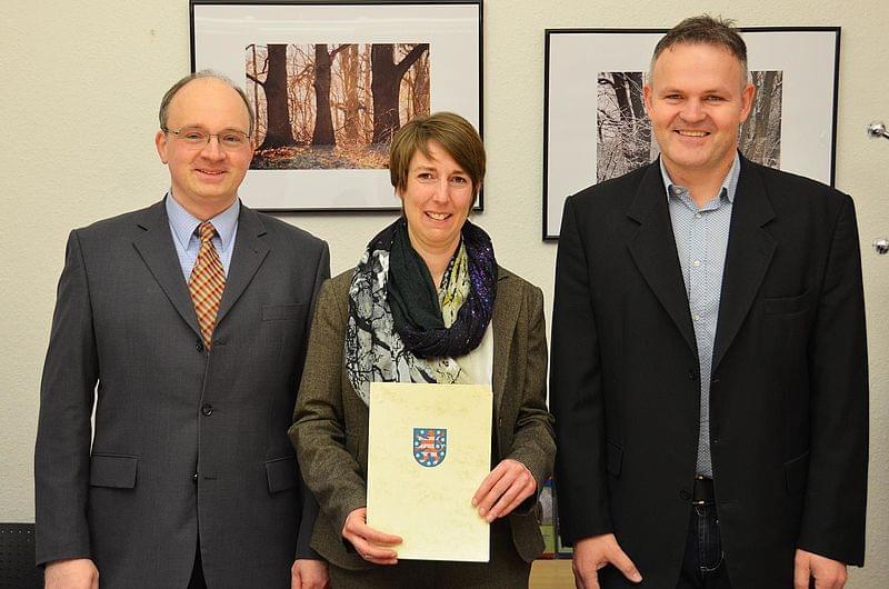 Prof. Dr. Gebhard steht zwischen Prof. Zahradnik und Prof. Wagner. Sie hält ihre Urkunde in den Händen. Im Hintergrund hängen zwei gerahmte Fotografien an der Wand.