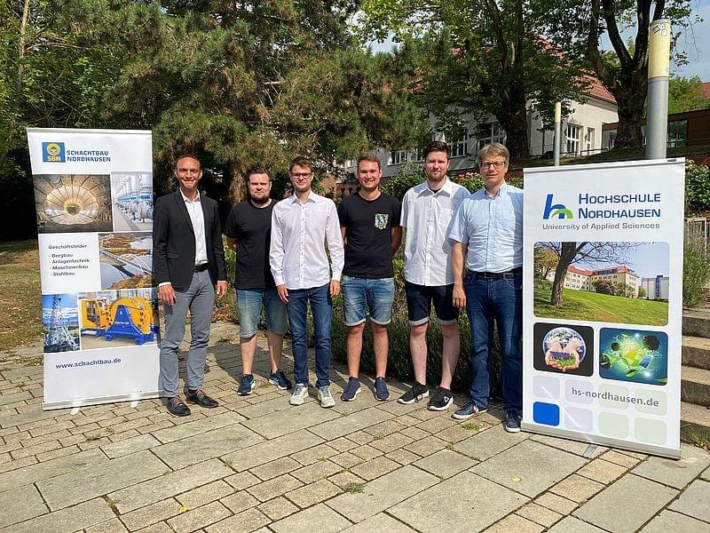 Teilnehmende stehen nebeneinander zwischen zwei Info-Rollups. Im Hintergrund ein Gebäude der Hochschule und Bäume.