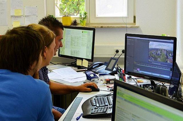 Three people are sitting in an office in front of several computer monitors. They are working together on a project, with a video or image editing programme open on one of the screens. The room is equipped with various office materials and papers, and daylight is streaming in through the window in the background.