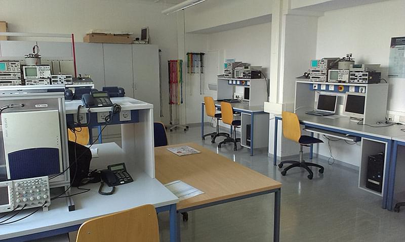 The picture shows several workstations with desks and chairs. On the desks are various technical devices, including oscilloscopes, spectrum analysers, power supply units and computers. Cabinets and shelves with other laboratory equipment can be seen in the background. The walls of the room are painted white and the windows let in daylight, which brightens up the room.