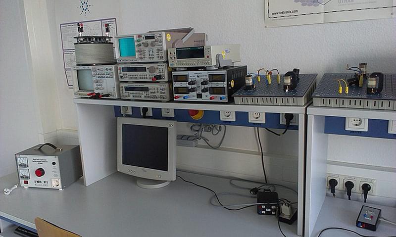 A communications laboratory with various technical devices and instruments. In the foreground is a computer monitor, with several measuring devices, oscilloscopes and power supply units stacked behind it. To the right are further devices and modules arranged on a table. The walls of the room are decorated with various posters and diagrams. The lab is bright and well lit, with a large window in the back that lets in natural light. There are several workstations with computers and chairs.