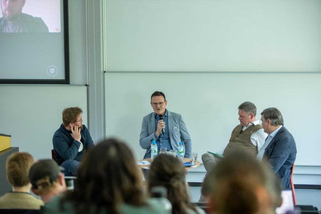 A panel discussion in a seminar room. Four men sit on chairs in front of an audience and discuss. Prof Jörg Wagner holds a microphone and speaks while the others listen attentively. In the background is a screen with a picture of another participant who is taking part in the discussion virtually. There are water bottles and glasses on the table in front of the speakers. The audience is partially visible.