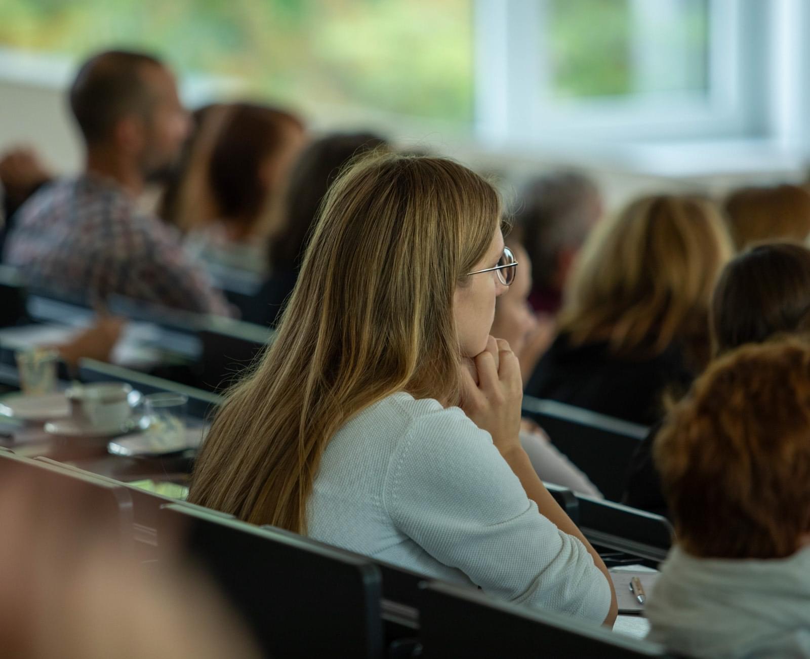 Studierende im Hörsaal. Eine junge Frau mit langen Haaren schaut nach vorne.