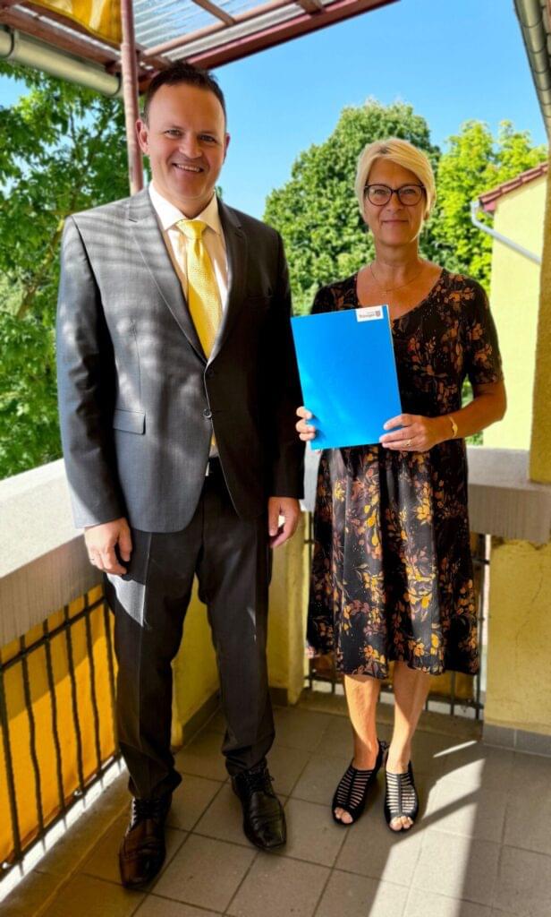 Das Bild zeigt zwei Personen, die nebeneinander auf einem sonnenbeschienenen Balkon oder einer Terrasse stehen und in die Kamera lächeln. Der Mann links trägt einen dunklen grauen Anzug mit einer gelben Krawatte, während die Frau rechts ein schwarzes, geblümtes Kleid trägt und eine blaue Mappe in den Händen hält. Im Hintergrund sind Grünpflanzen und ein Teil des Gebäudes zu sehen.