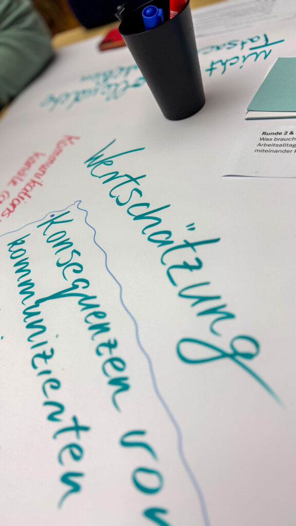 A close-up of a World Café table with a piece of paper on which terms such as "appreciation" and "communicate consistently" are written in green and red. Pens in a black cup can be seen in the background.