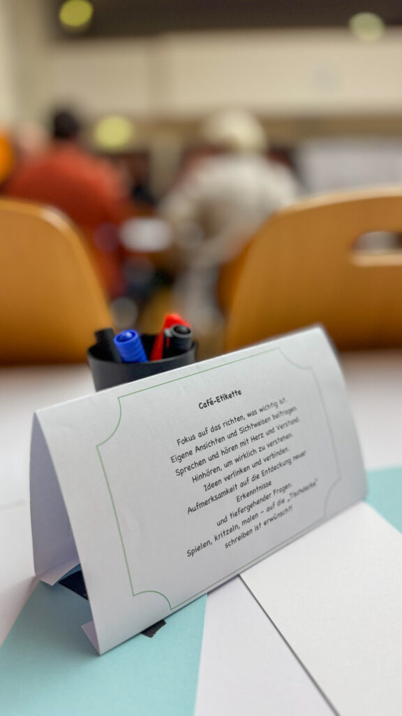 A table in an event room with a card on which the "café etiquette" for the World Café is described. In the background, several people are sitting on chairs in a blur, apparently taking part in discussions. A pencil cup with red and blue pens is also placed on the table.