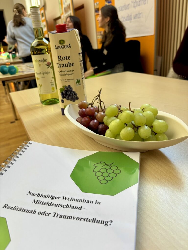 Ein Tisch mit einer Flasche Weißwein, einer Schale mit grünen Trauben und einem Heft mit der Aufschrift „Nachhaltiger Weinbau in Mähren/Piemont – Realität oder Illusion?