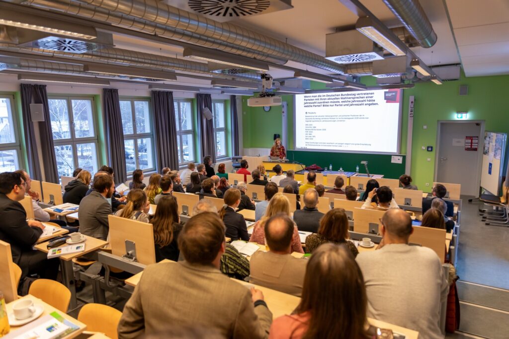 Ein Vortragsraum mit vielen Zuhörern, die in Reihen sitzen. Vorne hält Dr. Mirjam Jenny eine Keynote-Präsentation. Eine große Leinwand zeigt eine Folie mit Inhalten. Der Raum hat große Fenster, grüne Wände und eine moderne Beleuchtung.