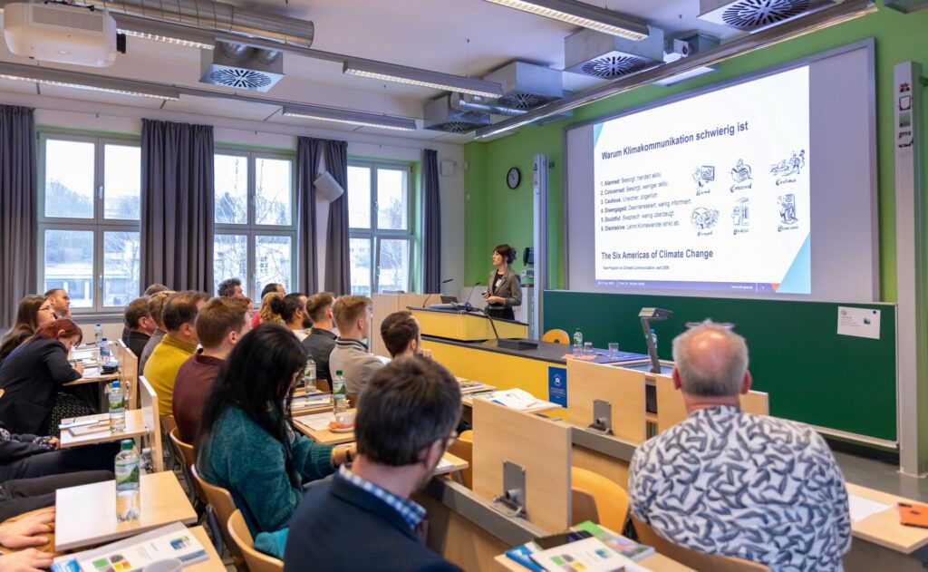 Ein Vortragsraum mit Teilnehmern, die an langen Tischen sitzen und Notizen machen. Prof. Dr. Nicole Harth hält eine Keynote-Präsentation und steht vor einer großen Leinwand mit einer Folienpräsentation. Der Raum hat große Fenster, grüne Wände und eine moderne Ausstattung.