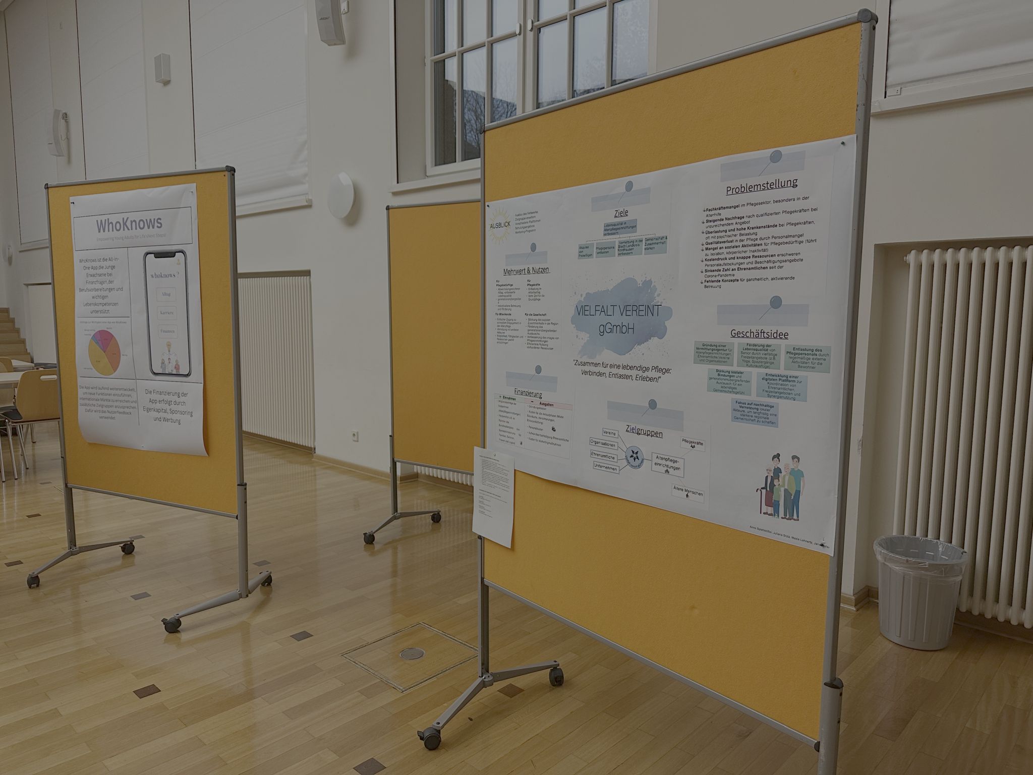 Several presentation walls with scientific posters stand in a large, bright room with a wooden floor and white walls. In the foreground is a poster entitled 'VIELFALT VEREINT gGmbH', which deals with the care and integration of different target groups. Next to it is another poster entitled 'WhoKnows', which describes an app for imparting knowledge. The presentation walls are mobile and have a yellow background. There are chairs in the background and a rubbish bin is placed on the wall.