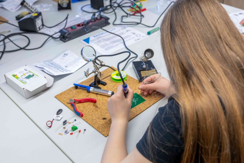 A person is soldering a green circuit board on a table, surrounded by tools such as pliers, wire and electronic components. A magnifying glass holder and other materials are within easy reach on the work surface,