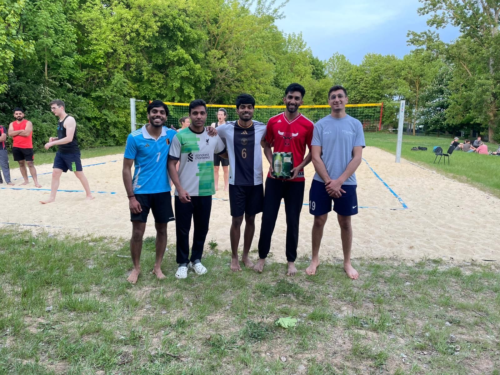 Five students, some in sports shirts, proudly hold a drinks bottle as a prize. They are standing on the university's sand court, with other teams in the background.