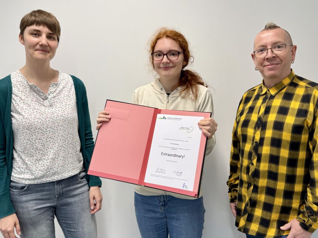 Drei Personen stehen vor einer weißen Wand, die mittlere Person hält eine aufgeschlagene rote Mappe mit einer Urkunde hoch. Der Titel auf der Urkunde lautet „Extraordinary!“ und stammt von der Hochschule Nordhausen. Alle drei blicken freundlich in die Kamera.