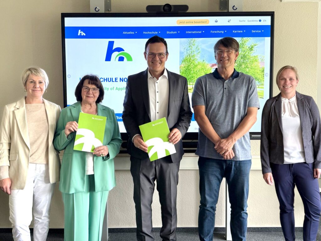 Five people stand next to each other in front of a screen with the Nordhausen University of Applied Sciences website. Two of them are holding green folders with the university logo. They are all smiling at the camera.