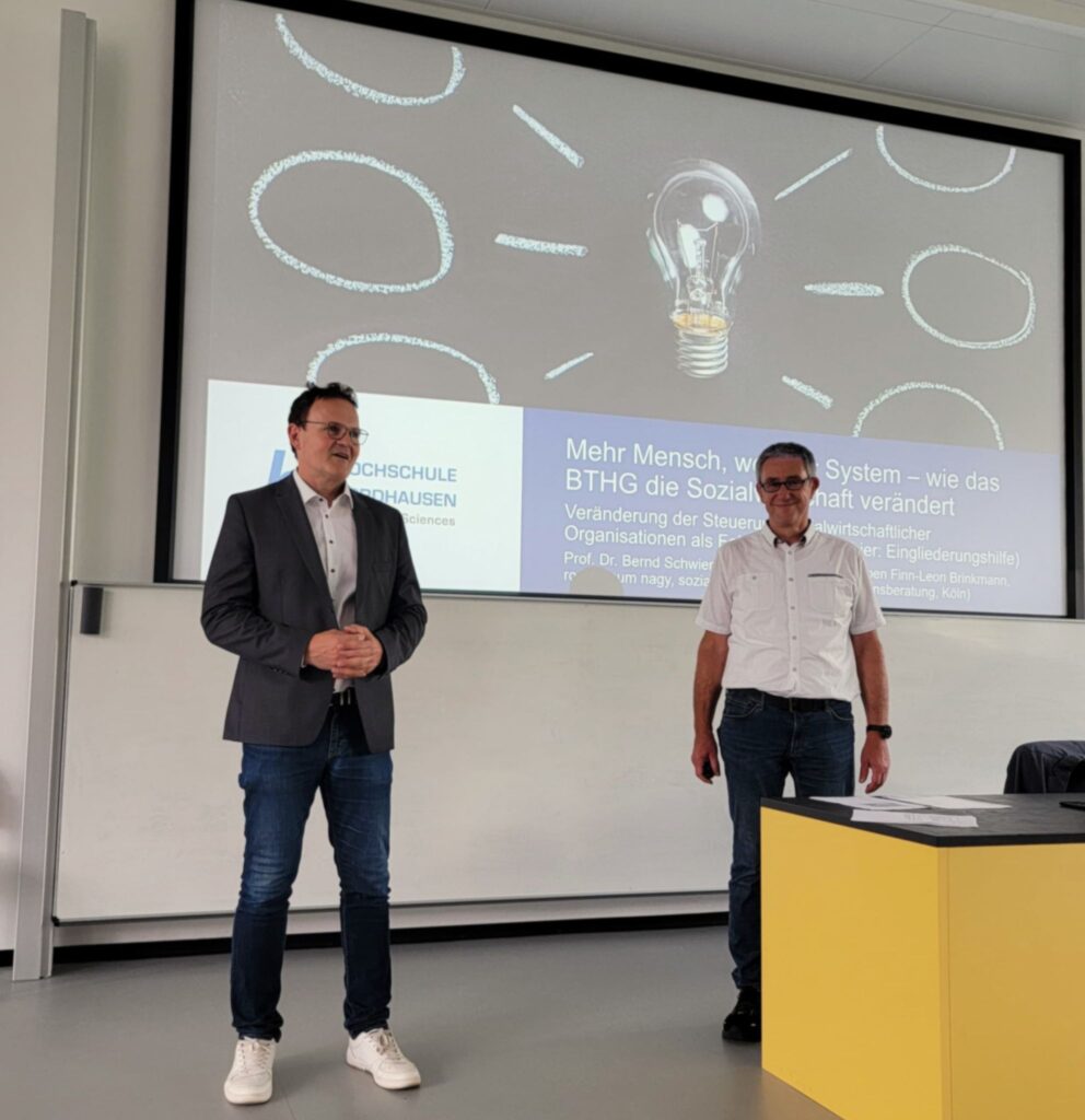 Two men, President Prof Dr Jörg Wagner on the left and Prof Dr Bernd Schwien on the right, stand in front of a presentation slide in the lecture theatre. The slide is entitled "More people, less system - how the BTHG is changing the social economy". Both appear to be in a good mood.