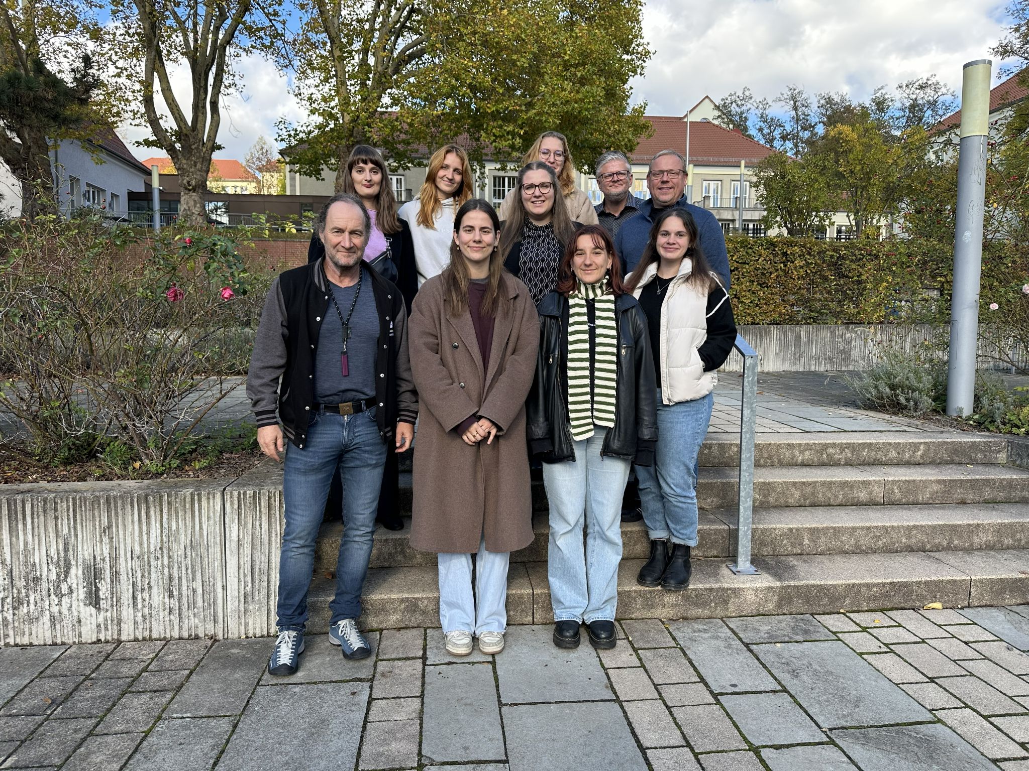 Group photo with ten people, taken outdoors on a staircase in front of autumn-coloured trees and buildings with red roofs. Five people are standing in the first row, including three women and two men; five more people are standing in the second row. They are all smiling at the camera, the atmosphere is friendly and relaxed.