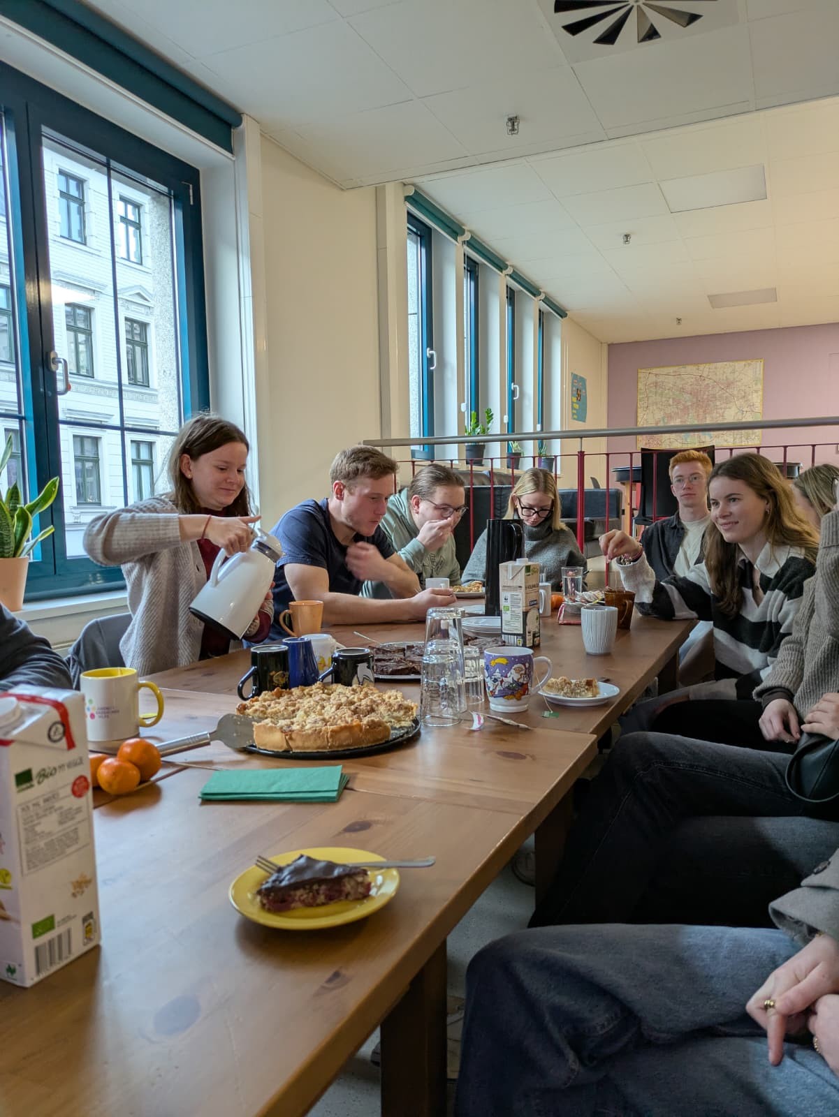 Eine Gruppe von Studierenden sitzt an einem langen Tisch mit Kuchen, Getränken und Geschirr und frühstückt gemeinsam in einem lichtdurchfluteten Raum. Eine Person schenkt Tee oder Kaffee aus einem Wasserkocher ein. Im Hintergrund sind große Fenster und eine Weltkarte an der Wand zu sehen.