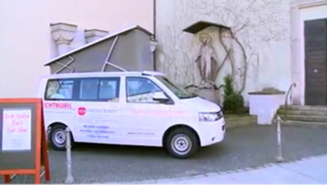 White confession mobile with the inscription „Oasenmobil - Zeit für dich“ (Oasis mobile - time for you) from the aid organisation „Kirche in Not“ (Church in Need) parked in front of a church next to a statue; in the foreground a display with the text „Zeit haben. Time for you.“