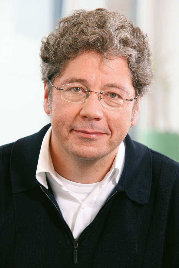 A man with curly grey hair and glasses, dressed in a white shirt and a dark jumper. He looks friendly into the camera and is part of the team of authors of the marketing textbook.