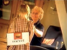 The picture captures a heart-warming moment in warm golden tones. A young girl with pigtails and a chequered dress stands with her back to the viewer and holds a Merci packet behind her back or is about to hand it over. Her counterpart is an older woman with blonde hair who smiles lovingly and joyfully at the child. The atmosphere is homely and familiar.