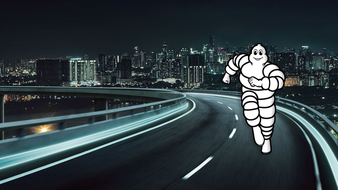 The graphic shows a modern motorway bridge at night, whose road markings appear as bright strips of light due to long exposure. In the right foreground, the iconic advertising figure „Bibendum“ (the Michelin man) runs towards the viewer with a friendly smile. In the background, the brightly lit skyline of a large city can be seen at night.