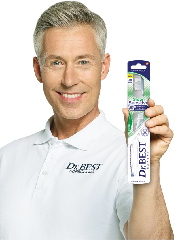 The picture shows a portrait of a grey-haired man wearing a white polo shirt with the inscription „Dr.BEST FORSCHUNG“. He smiles at the camera and holds up a toothbrush in its original packaging in his right hand. The packaging is labelled „Green Sensitive“ and „EXTRA SOFT“. A seal on the pack identifies the product as „CLIMATE NEUTRAL“.