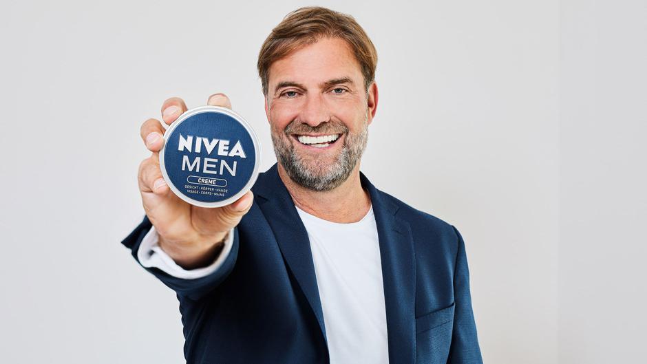 The picture shows Jürgen Klopp in a dark blue jacket over a white T-shirt against a light grey background. He is smiling at the camera and holding a blue can of „NIVEA MEN CREME“ in his outstretched right hand. The can is positioned in such a way that the product name is central and clearly legible for the viewer.