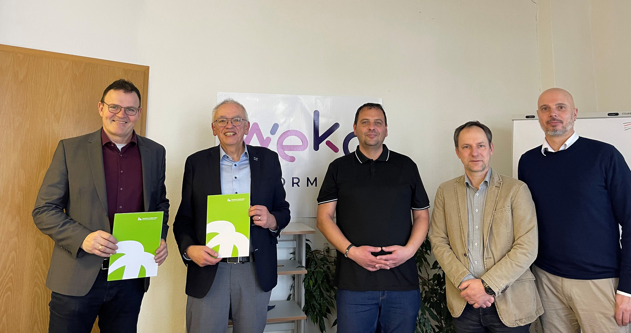 Fünf Männer stehen nebeneinander in einem Büro vor einem WEKO Informatik Banner. Zwei von ihnen halten grüne Mappen der Hochschule Nordhausen in der Hand. Das Foto dokumentiert die besiegelte Zusammenarbeit zur Fachkräftesicherung.