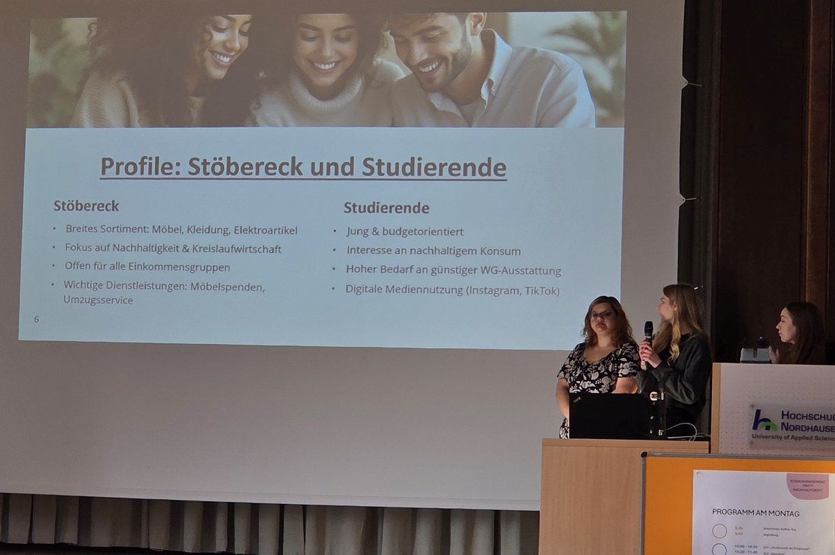 Drei junge Frauen präsentieren vor einer Folie mit den Profilen „Stöbereck“ und „Studierende“, auf der Themen wie Budgetorientierung, Nachhaltigkeit und Möbelspenden beschrieben sind.