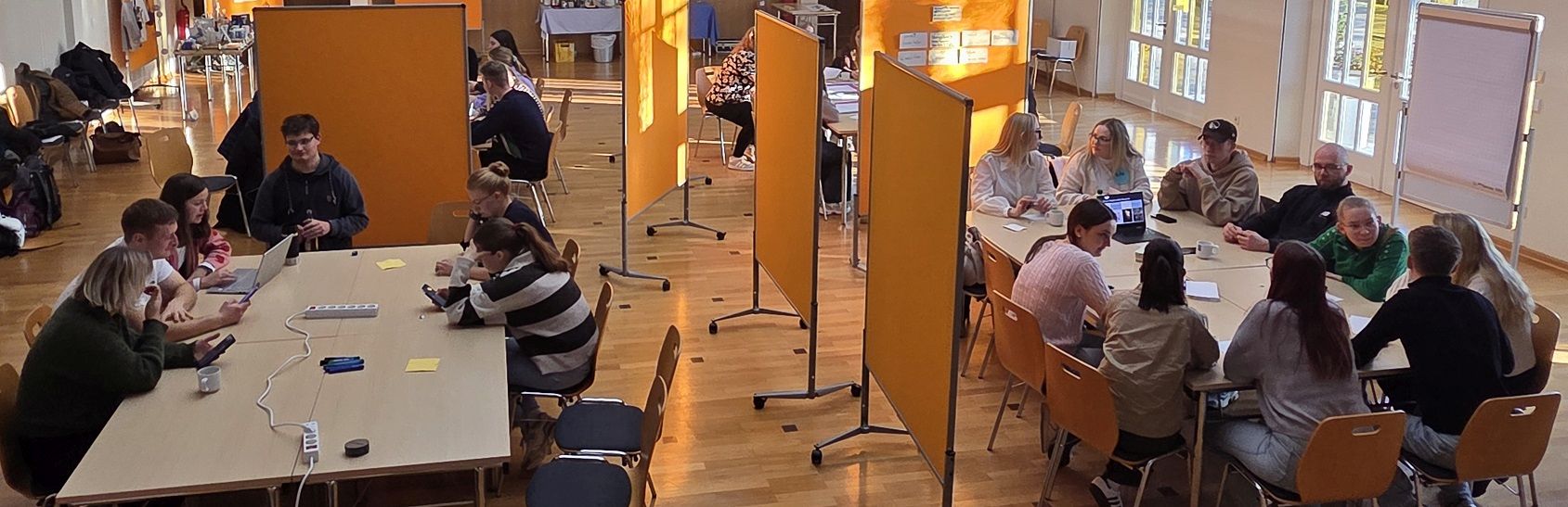 Mehrere Gruppen junger Menschen arbeiten an langen Tischen in einem hellen Raum mit mobilen Pinnwänden. Laptops, Stifte und Notizzettel sind auf den Tischen verteilt.