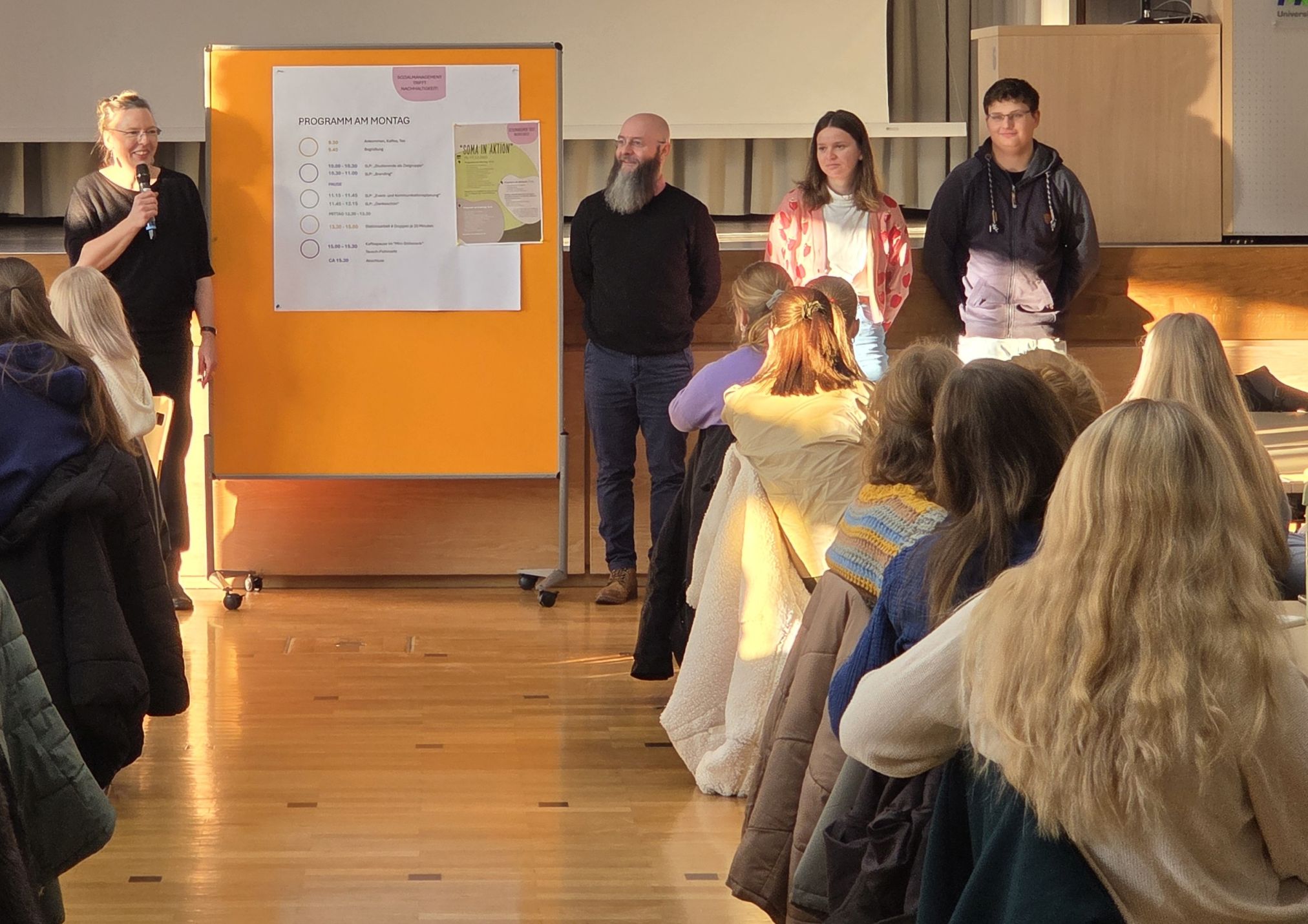 Vier Personen stehen vor einer Gruppe sitzender Teilnehmerinnen. Links wird ein Programmplakat mit dem Tagesablauf gezeigt. Eine Frau spricht in ein Mikrofon.