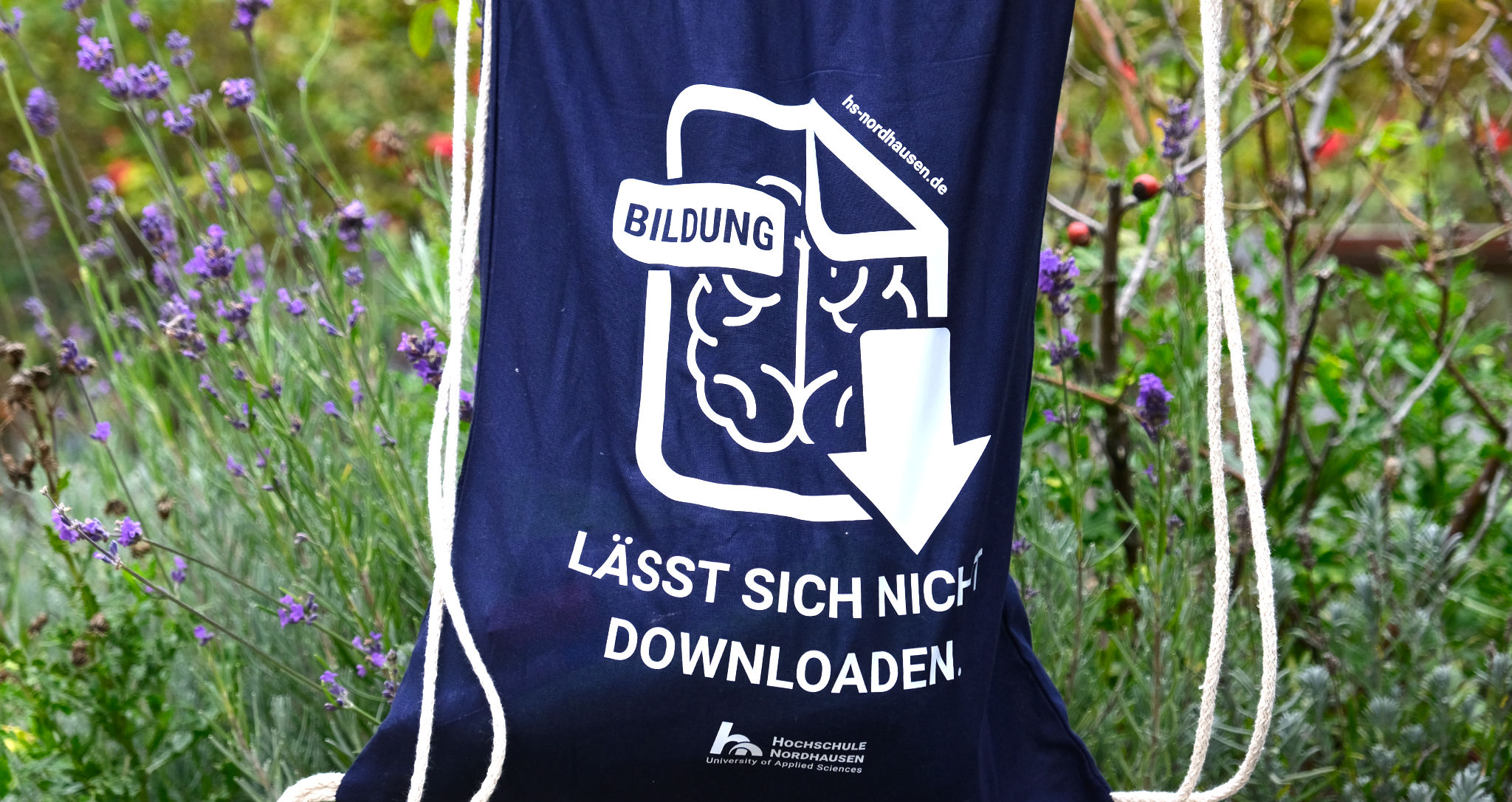 A blue gym bag with the inscription "Education cannot be downloaded". Underneath is the logo of Nordhausen University of Applied Sciences. The green campus in the background.